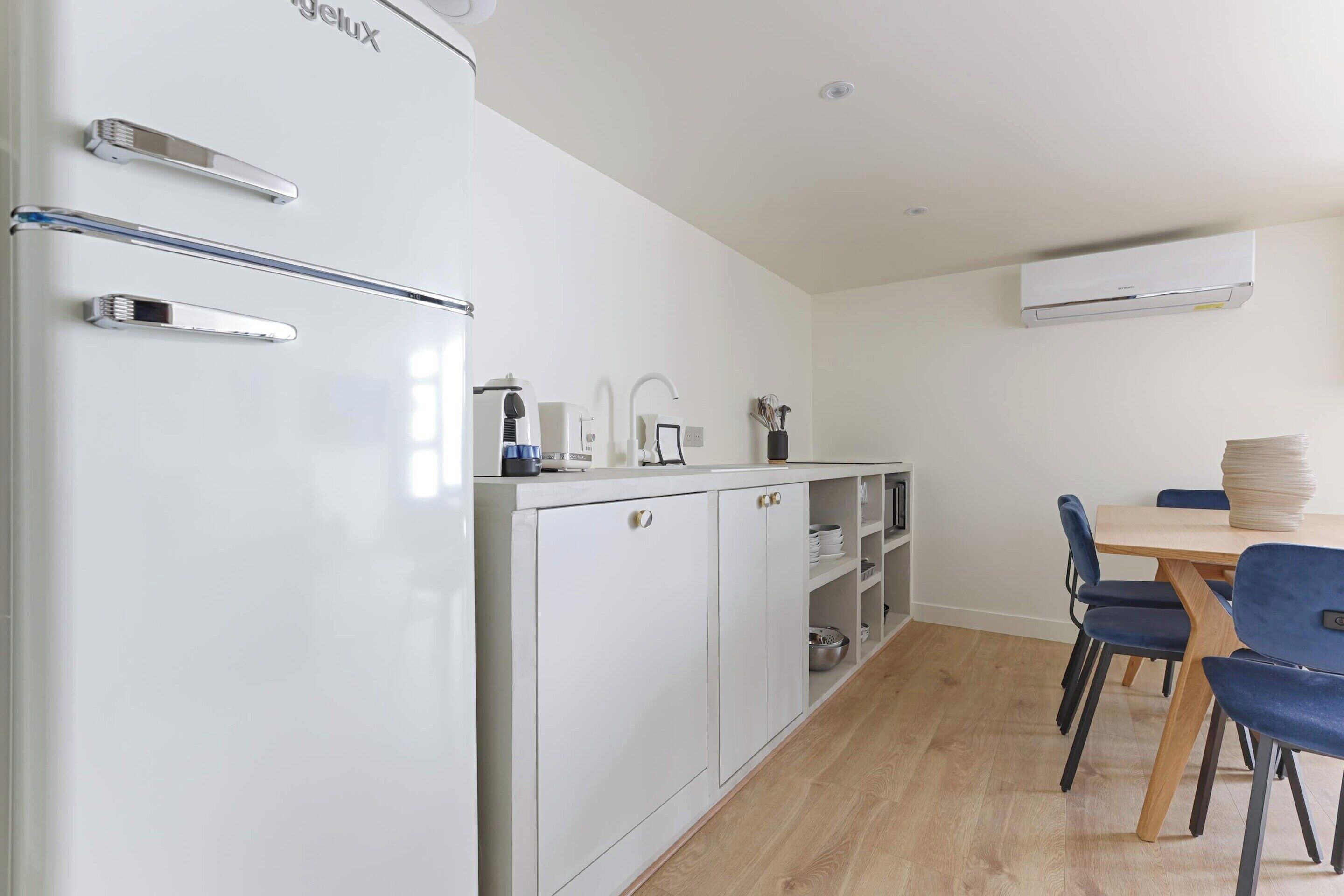 A compact kitchen with a white refrigerator, a microwave, and a sink. There is a dining table with chairs and a stack of plates on the table.