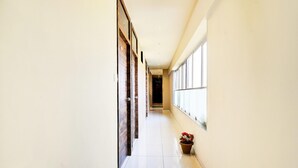A long, narrow hallway with a tiled floor, wooden doors on one side, and large windows on the other. - Super Hotel O Somnath Formerly Hotel Somnath Tirth (Veraval)