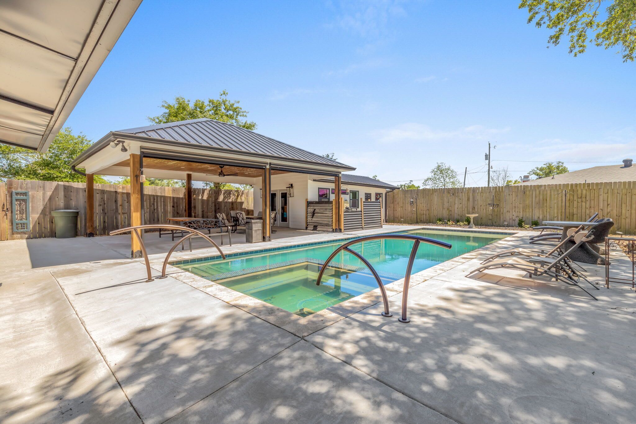 A backyard with a swimming pool, a covered patio, and outdoor furniture.