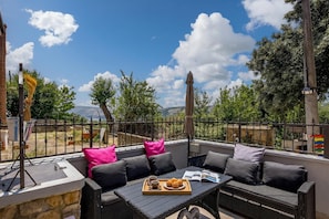 An outdoor seating area with a sofa and table, a tray with muffins and a book, and a view of trees and mountains.