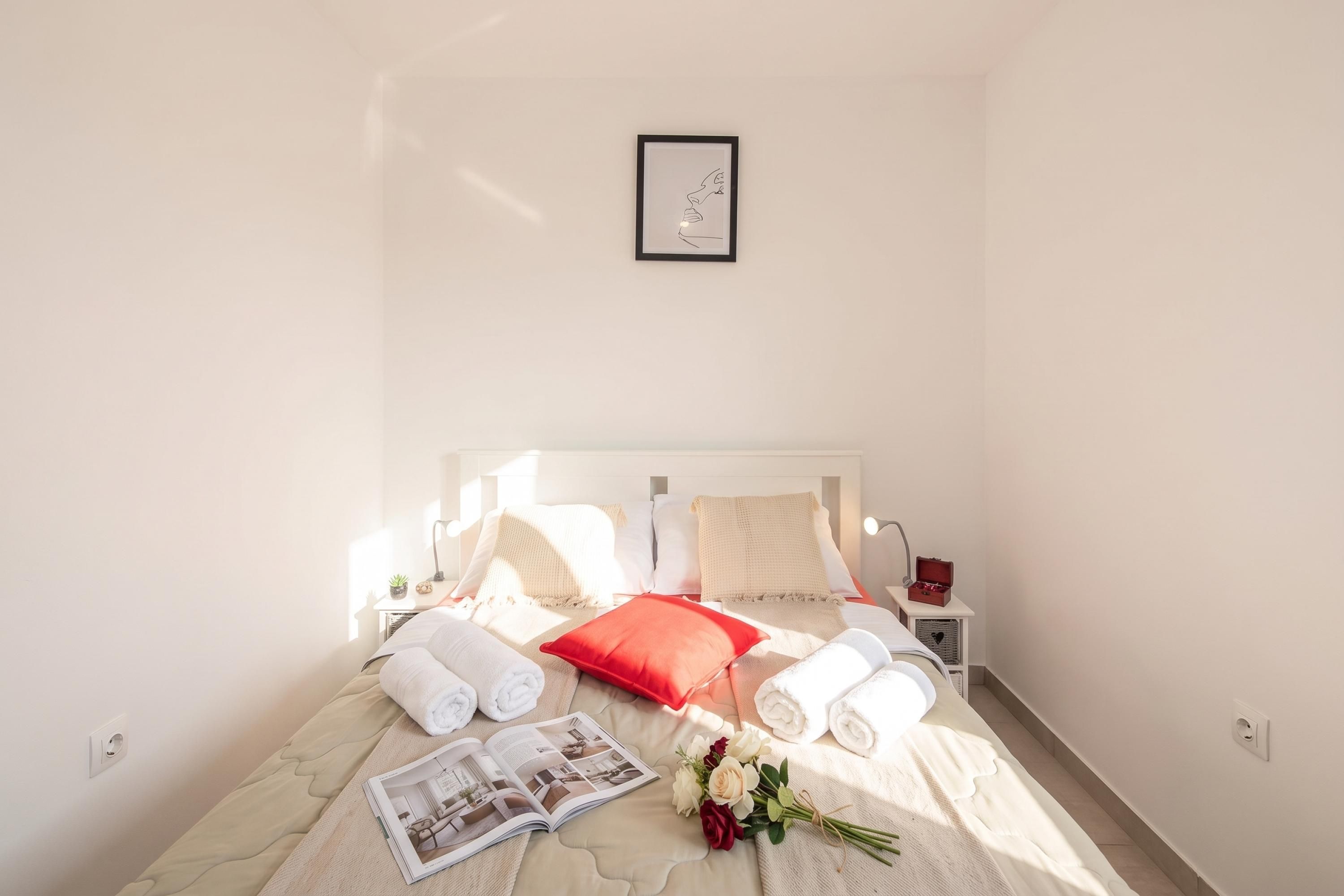 A neatly made bed with white and red pillows, a bedside table with a lamp, and a framed picture on the wall.