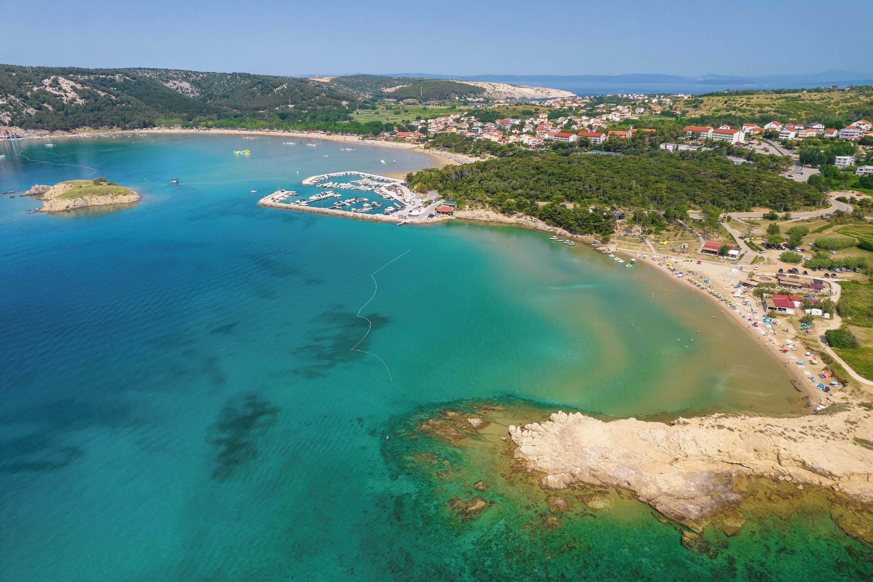 An aerial view of a coastal area with clear waters, a marina, and a residential area with houses and greenery.