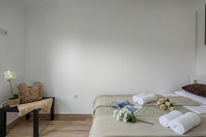 Siististi järjestetty makuuhuone, jossa on sänky, pieni pöytä, jossa on kori ja ruukkukasvi, sekä yöpöytä, jossa on rullalle taiteltuja pyyhkeitä ja kukkia.