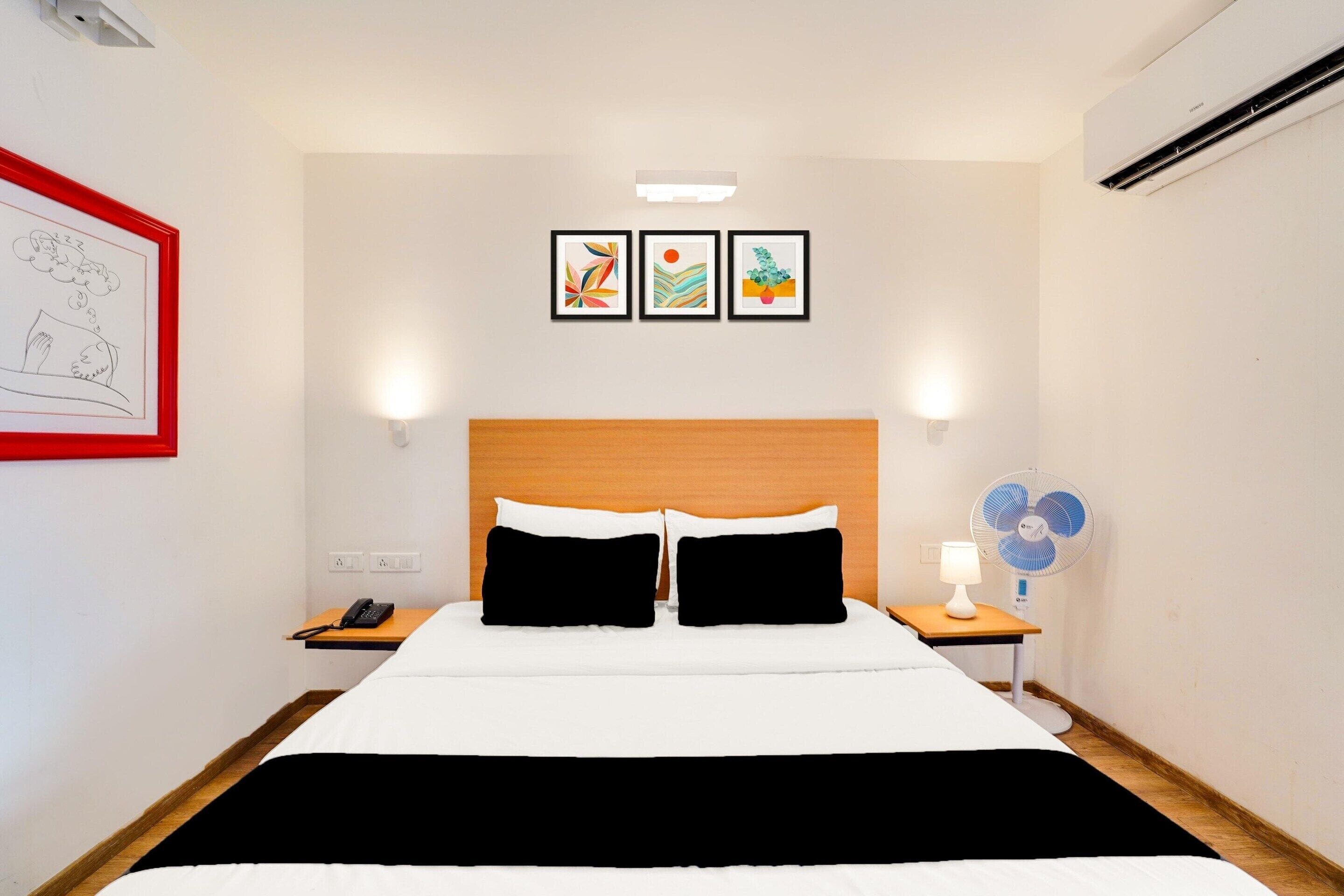 A hotel room with a bed, bedside tables, a wall-mounted phone, a fan, and framed artwork on the wall.
