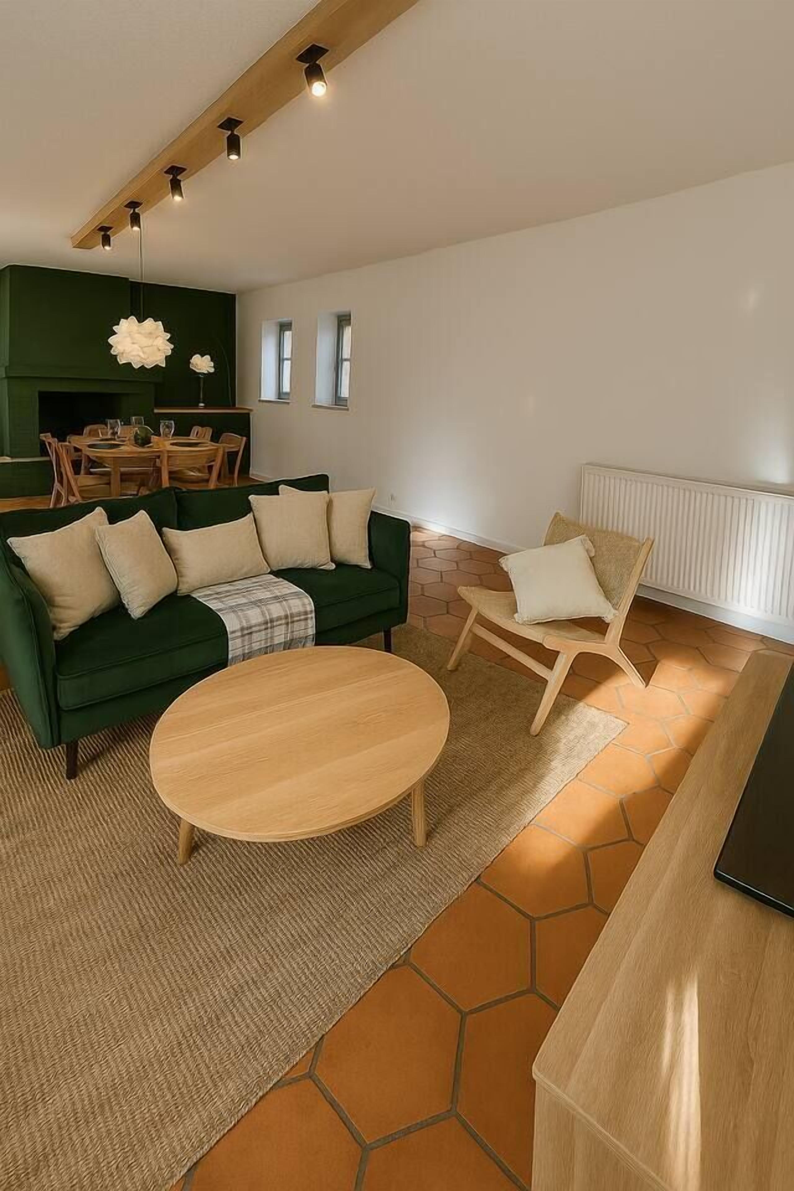 Un salon moderne avec un canapé vert, une table basse en bois et un fauteuil de couleur claire. On aperçoit à l’arrière-plan un coin repas avec une table et des chaises en bois.