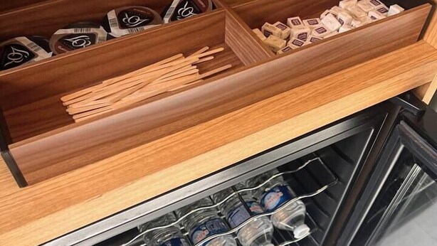 Une cuisine avec des tiroirs en bois contenant des sachets de thé et des ustensiles de cuisine en bois, ainsi qu’un réfrigérateur rempli de bouteilles d’eau.