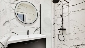 Un baño con pared de mármol blanco, un espejo redondo, un mueble negro y una ducha con cabezal de ducha manual.