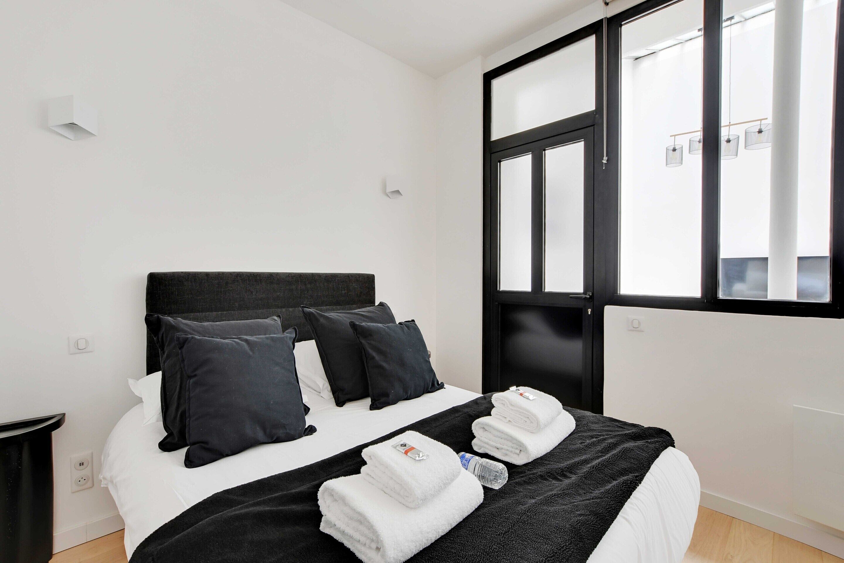A hotel room with a large bed, black headboard, white bedding, and a window with blinds.