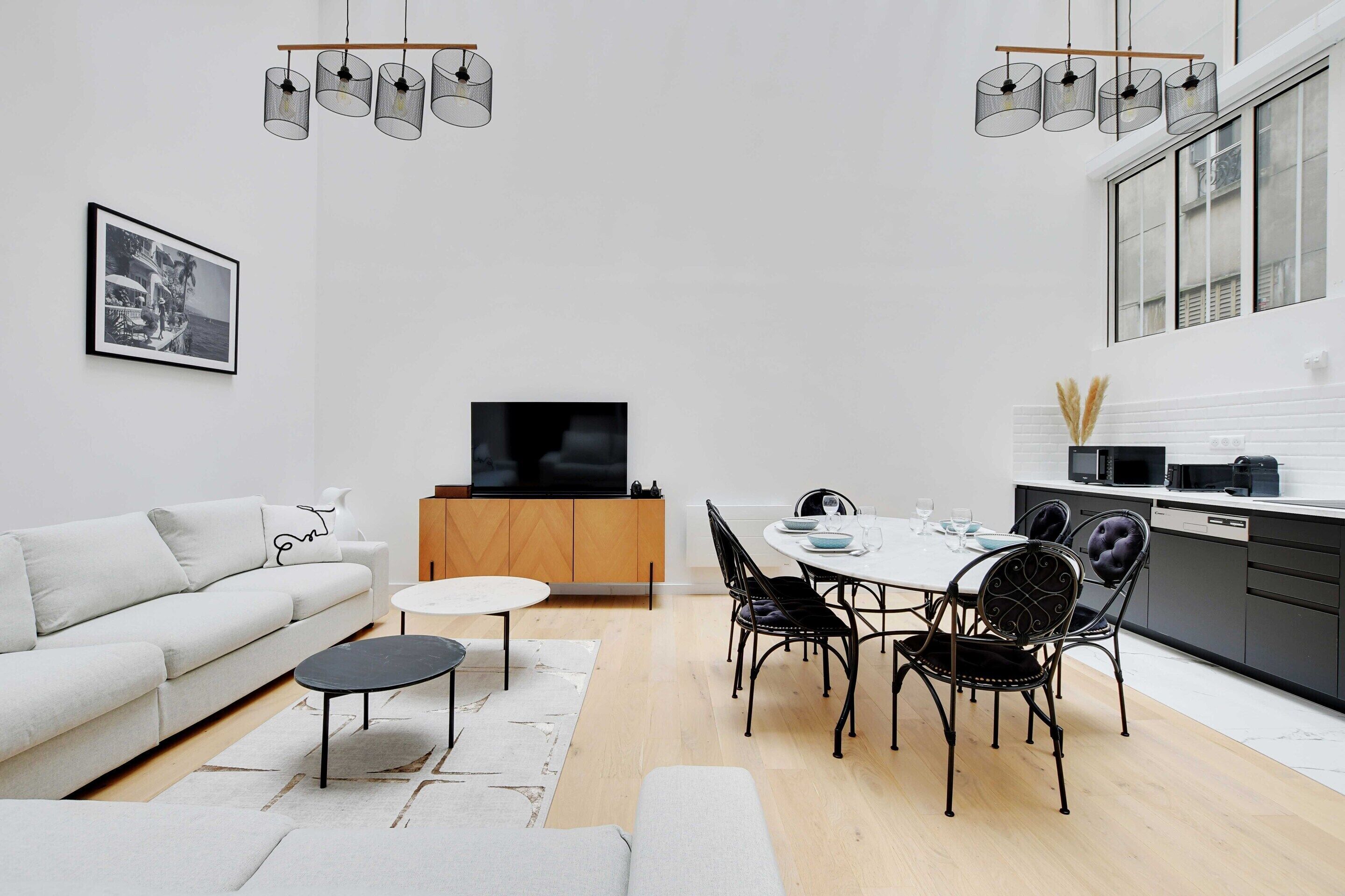 Ein modernes Wohnzimmer mit Sofa, Esstisch mit Stühlen, Fernseher und Küchenzeile mit Schränken und Geräten.