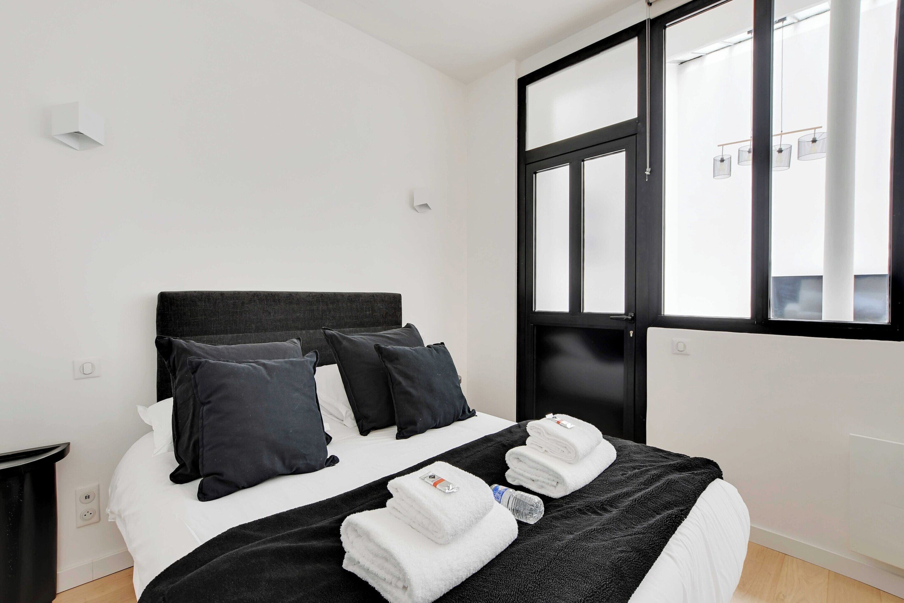 Habitación de hotel con cama, cabecero negro, ropa de cama blanca y una ventana con persianas.