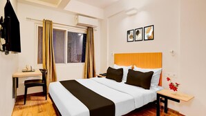 A hotel room with a bed, a desk, a chair, a window with curtains, a bedside table with a vase of flowers, and three framed pictures on the wall. - Spacious Comfort Stays Near Aerospace Museum (Bengaluru)