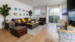 Un salon spacieux avec un canapé en cuir marron, un fauteuil blanc et une table basse. Des cadres photos sont accrochés aux murs et un ventilateur de plafond est installé.