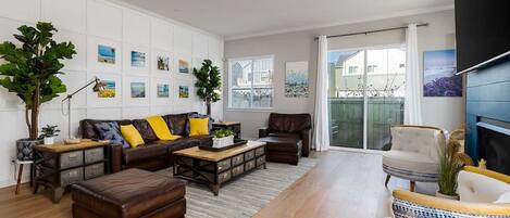 A spacious living room with a brown leather sofa, a white armchair, and a coffee table. There are wall-mounted picture frames and a ceiling fan.