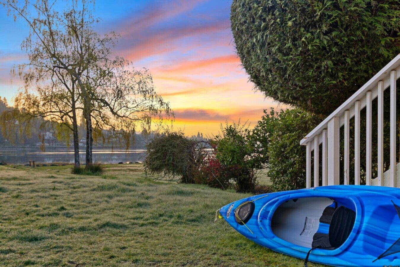 Een blauwe kajak op het gras, nabij een witte schutting, met bomen en een zonsondergang op de achtergrond.