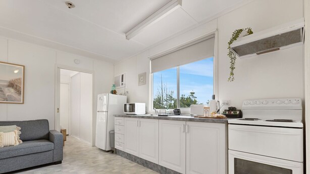 A modern kitchen with white cabinets, a grey sofa, and a window with a view of trees. - Bird and Beach: Close to walking trails leading to the beach and Lake Bunga (Lakes Entrance)