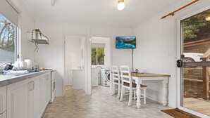 A modern kitchen with white cabinetry, a dining area with a wooden table and chairs, and a TV mounted on the wall. - Bird and Beach: Close to walking trails leading to the beach and Lake Bunga (Lakes Entrance)