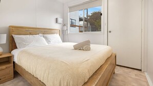 A bedroom with a wooden bed, white bedding, a bedside table with a lamp, and a window with blinds. - Bird and Beach: Close to walking trails leading to the beach and Lake Bunga (Lakes Entrance)