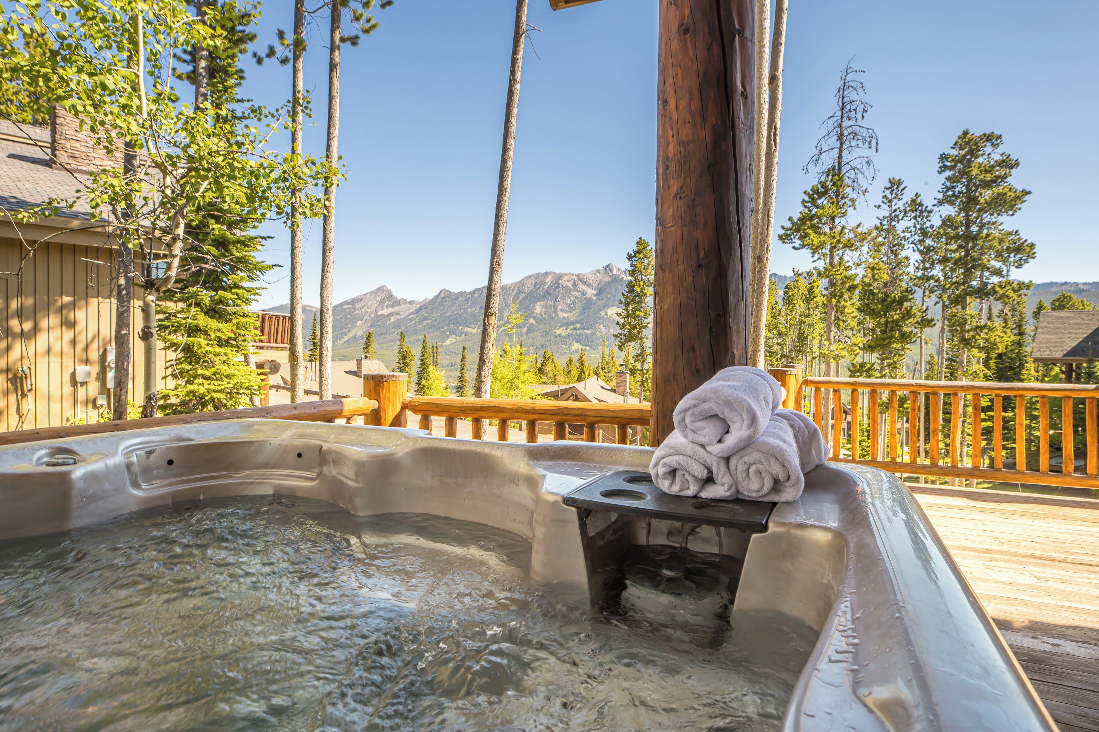 Un bain à remous avec une terrasse en bois, entouré d’arbres et avec des montagnes en arrière-plan.