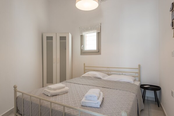 Una cama bien hecha con colcha acolchada y toallas dobladas, un armario blanco, una mesita redonda pequeña y una ventana con cortina blanca.