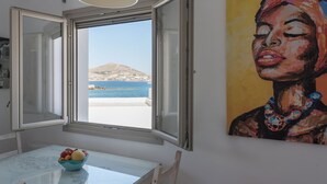 An open window with a view of a beach and mountain, a table with a bowl of fruit, and a painting on the wall. - Seaview House 2 From the Town (Paros)