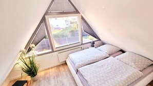 Ein Schlafzimmer mit schrägem Dach, einem Bett mit gemusterten Betttextilien, einem Heizkörper, einer Zimmerpflanze und einem kleinen Tisch.