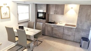Eine moderne Küche mit grauen Schränken, einem eingebauten Backofen und einem Spülbecken. Ein Esstisch mit Stühlen steht unter einem Fenster.