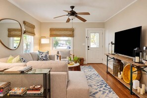 A living room with a sofa, coffee table, and a TV. There is a ceiling fan, a door, and a window with blinds.