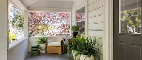 A covered porch with a wicker chair, potted plants, and a view of autumn trees.