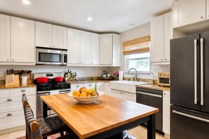 A modern kitchen with white cabinets, a stainless steel refrigerator, a microwave, and a wooden dining table with a bowl of fruit.