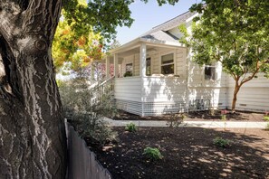 Una casa blanca con porche, un árbol en primer plano y un jardín con mantillo y plantas.