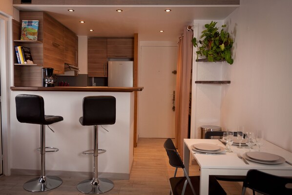 Una cocina compacta con armarios de madera, un refrigerador empotrado y una cafetera. Hay dos taburetes de bar y una mesa de comedor con platos y vasos.