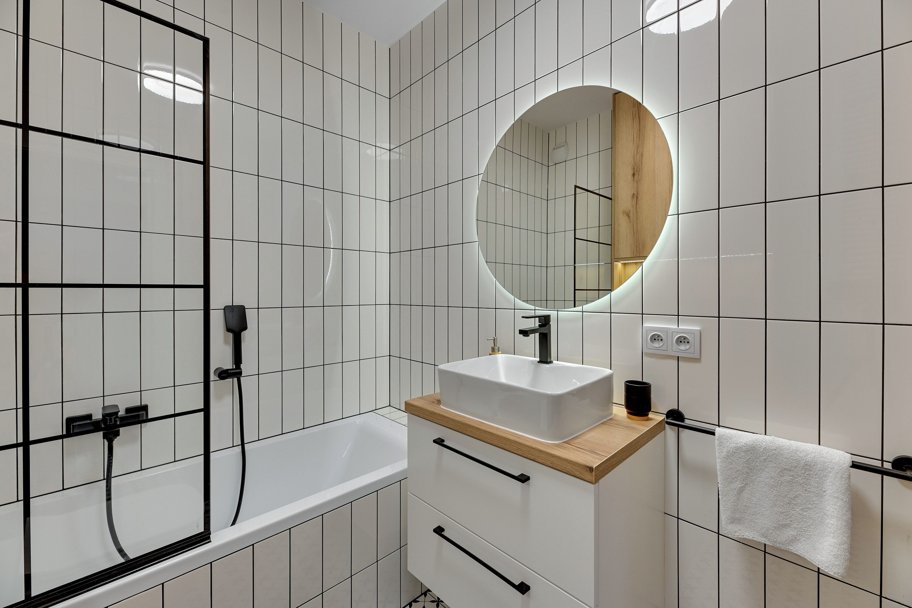Ein modernes Badezimmer mit weißen Fliesen, einem Holzwaschtisch, einem weißen Waschbecken, einem runden Spiegel, einer Badewanne und einem Handtuchhalter.