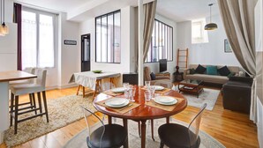 A modern living room with a dining area, large windows, and a sofa. - Lumunous apartment - 1BDR/5P au coeur du Marais (Paris)
