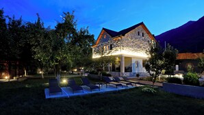 A stone house with a sloped roof, a swimming pool with lounge chairs, and a well-lit garden area. - Plis Bujtina (Margegaj)