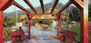 Una pérgola de madera con columpios, un patio con mesas y sillas, y vistas a un paisaje montañoso.