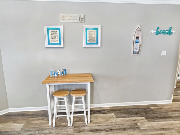 Una habitación con una mesa de madera y dos sillas blancas, un cuadro en la pared y un pequeño adorno con la palabra 'beach'.