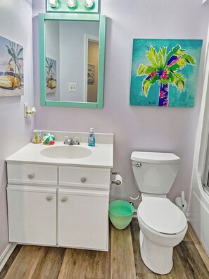 Un baño con lavamanos blanco, espejo e inodoro. Hay un balde verde junto al inodoro y un cuadro de una palmera en la pared.