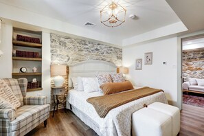 Un dormitorio con pared de piedra, una cama con ropa blanca, una mesita de noche con lámpara y una silla.