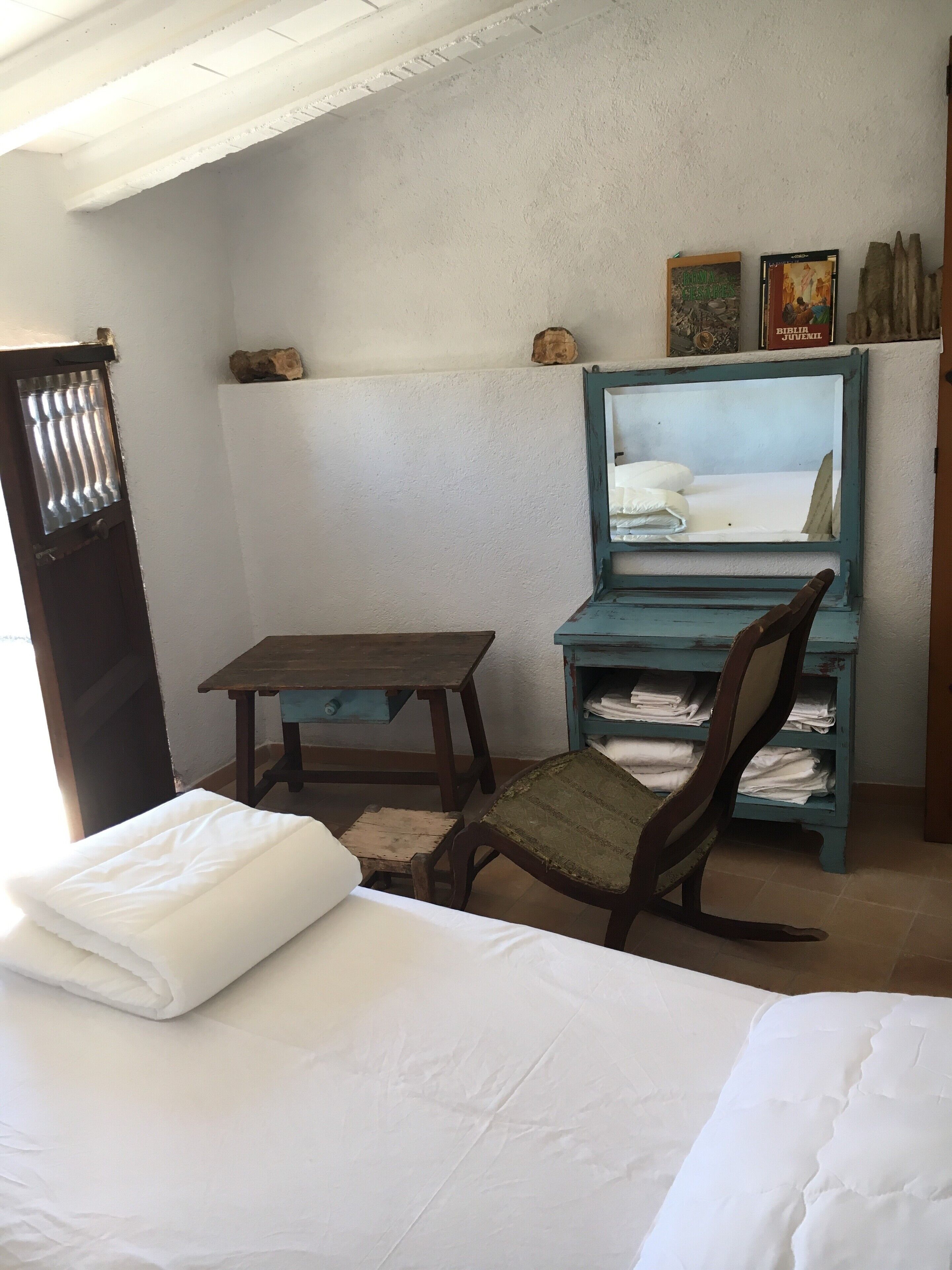 A room with a bed, a wooden table, a chair, a shelf with folded clothes, and a wall-mounted mirror.