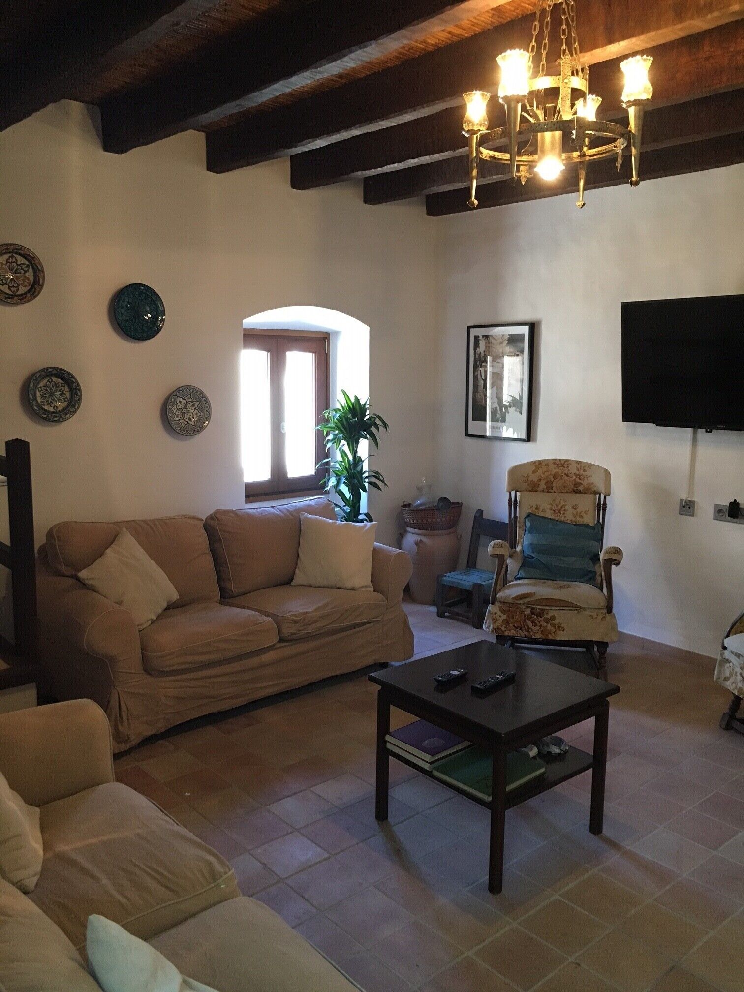 A living room with a sofa, armchair, and coffee table. There is a TV mounted on the wall and a chandelier hanging from the ceiling.