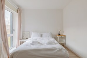 Una cama bien hecha con ropa blanca y toallas, una mesita de noche con lámpara y una ventana con cortinas.