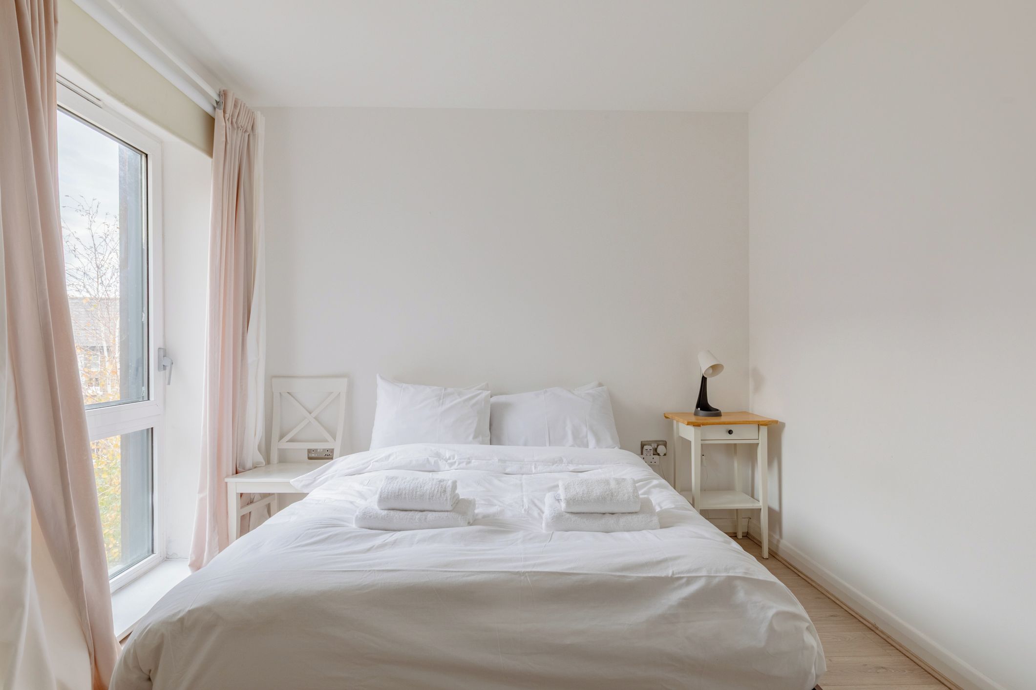 A neatly made bed with white linens and towels, a bedside table with a lamp, and a window with curtains.