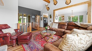 A spacious living room with a large brown leather sofa, a glass coffee table, and a red patterned rug. There are mounted deer heads on the wall, and a wooden ceiling beam is visible. - Mountain View, 3 minutes to downtown Banner Elk, easy access and ample parking!  (Beech Mountain)