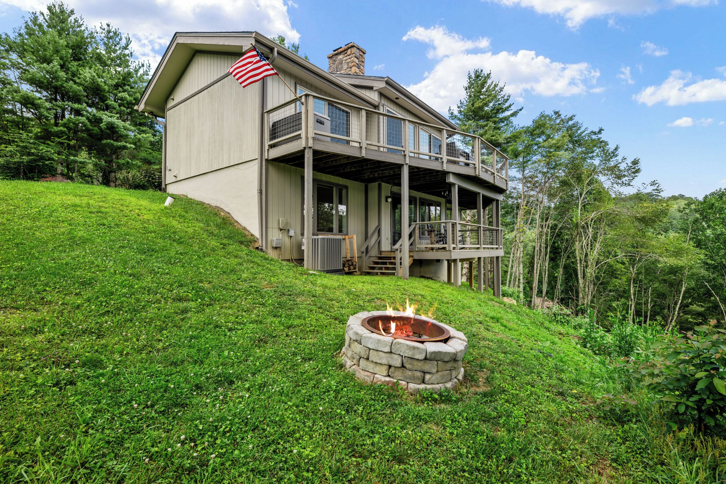 A two-story house with a stone fire pit in front, surrounded by lush greenery.