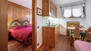 Una habitación compacta con cama de madera, una pequeña kitchenette, una zona de comedor con mesa y sillas, y una ventana con cortinas traslúcidas.