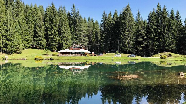 Un refugio junto al lago con una edificación grande rodeada de bosque denso, un reflejo claro en el agua y un pequeño muelle con barcos.