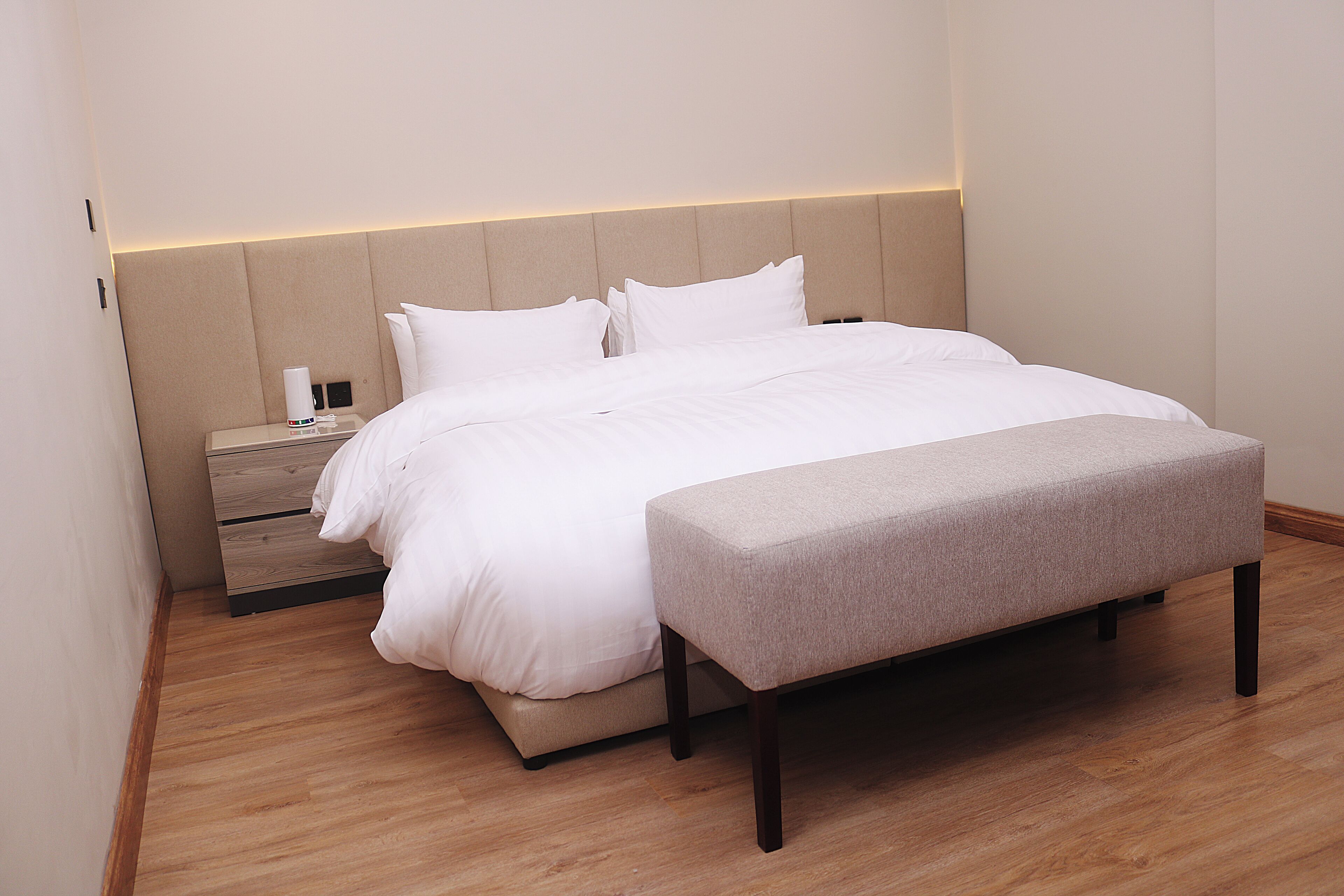 A modern hotel room with a large bed, a bedside table with a lamp, and a bench.