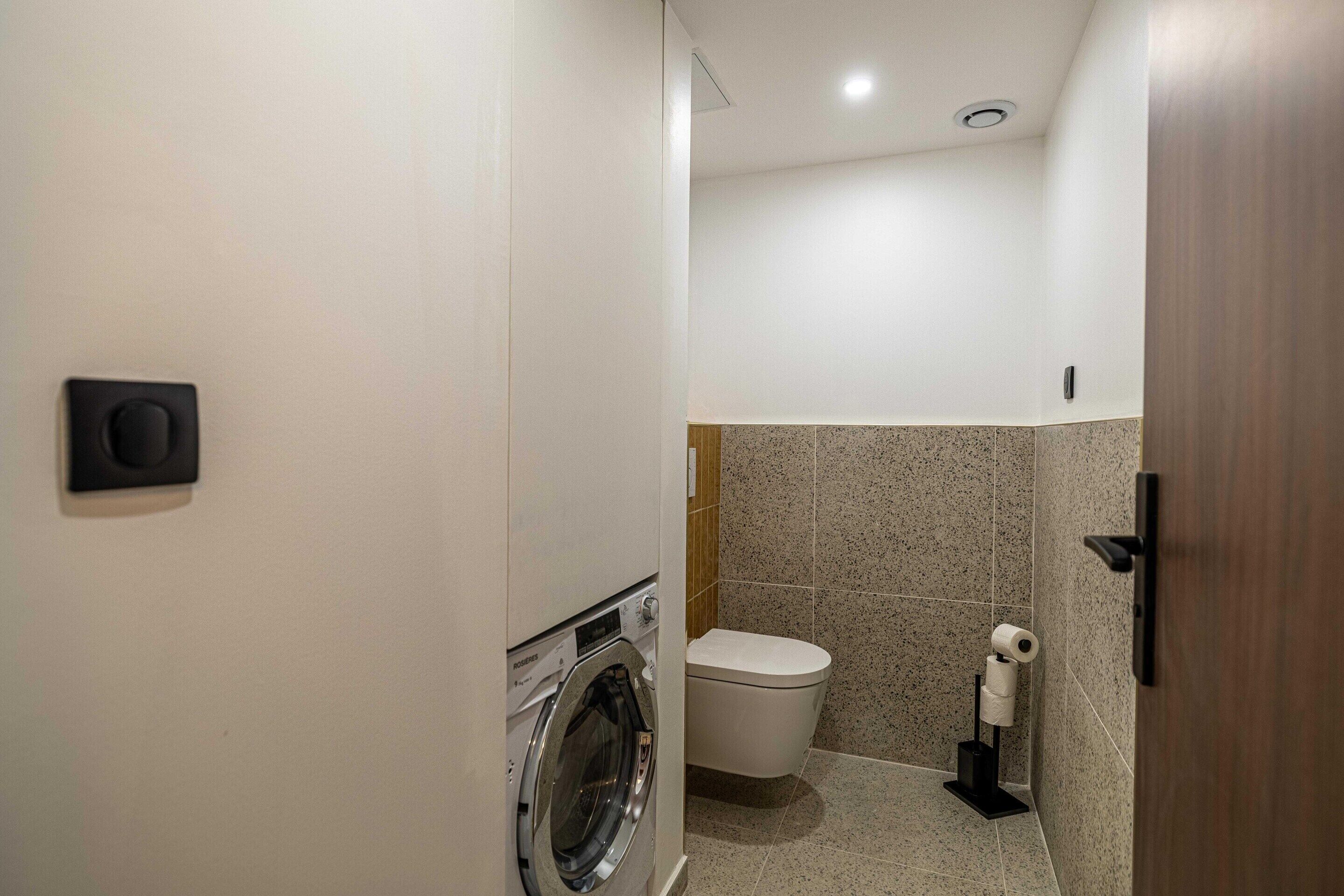 A compact bathroom with a washing machine, toilet, and a toilet paper holder.