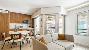 A modern kitchen with wooden cabinets, a dining area with a white table and chairs, and a living area with a beige sofa and a window with shutters. - Charming Flat - 2br-6p - Cannes (Cannes)