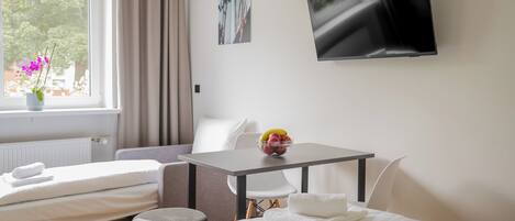 A hotel room with a bed, a small table with a bowl of fruit, a TV on the wall, and a window with curtains.